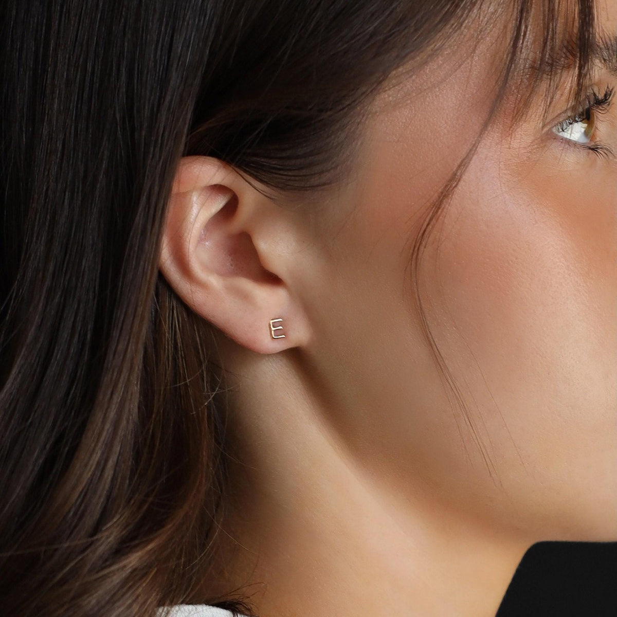 Close-up profile of a woman wearing a tiny gold letter E stud earring in her earlobe.