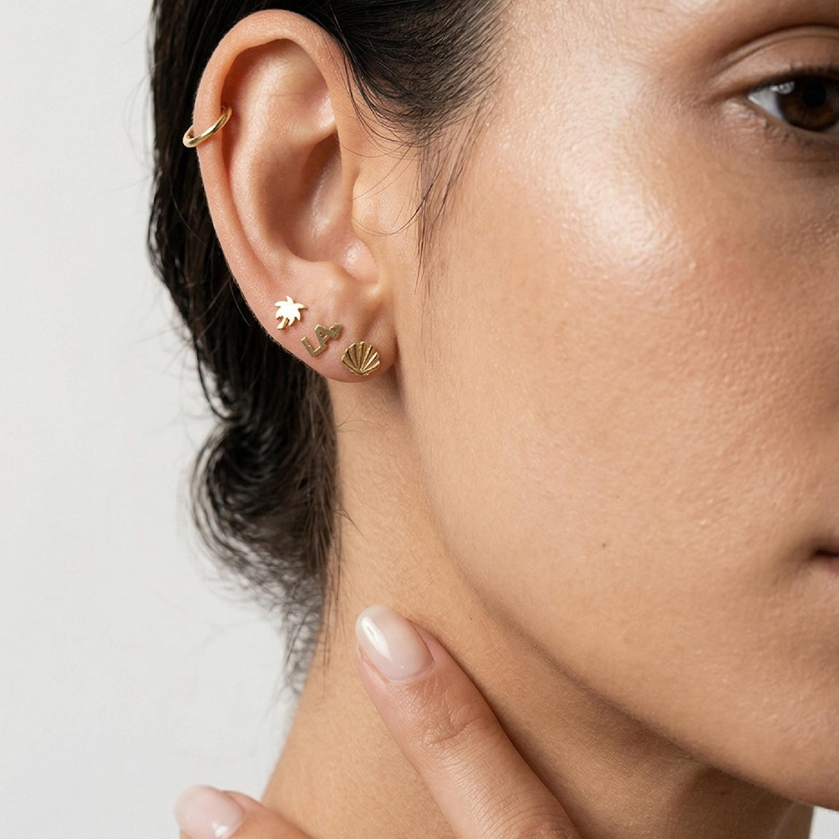 Close-up of a person ear wearing a small gold LA heart stud earring in the second lobe position, accompanied by a palm tree and seashell stud.