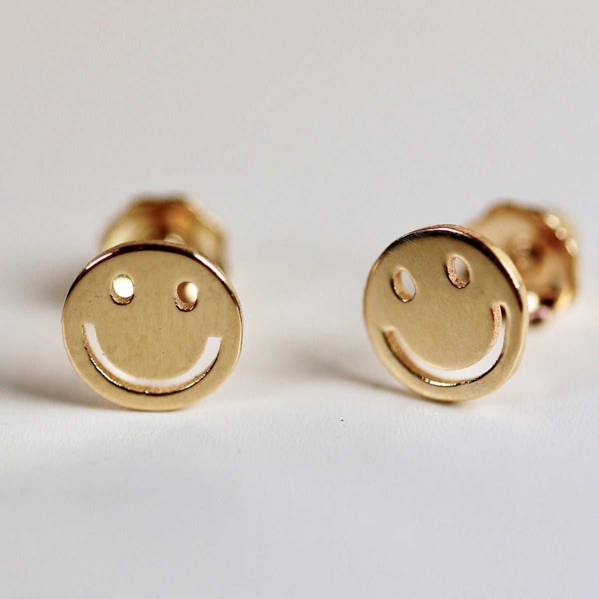 Two gold stud earrings shaped like smiling faces standing upright on a white surface.