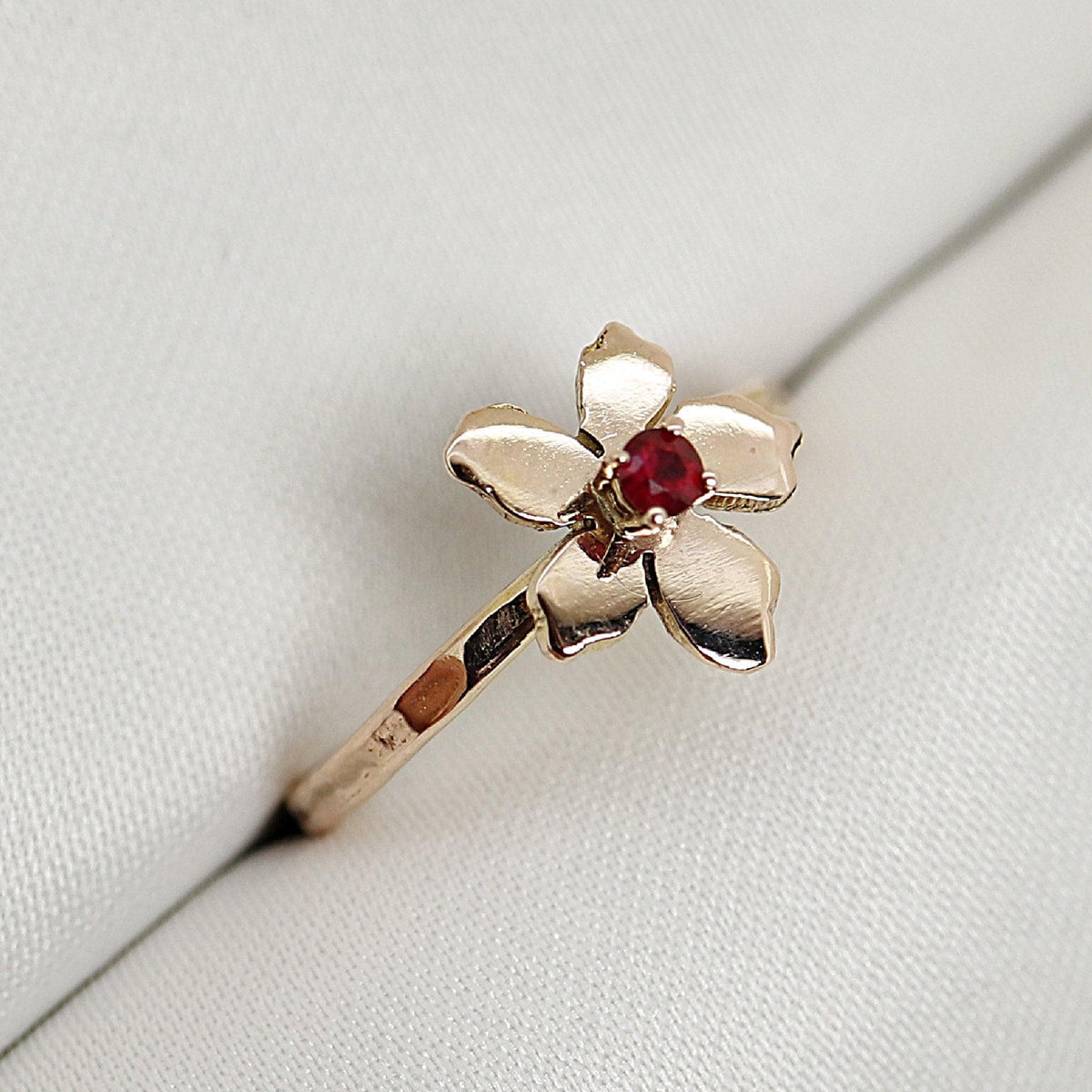 A gold flower ring with a red stone center rests on a white textured surface.