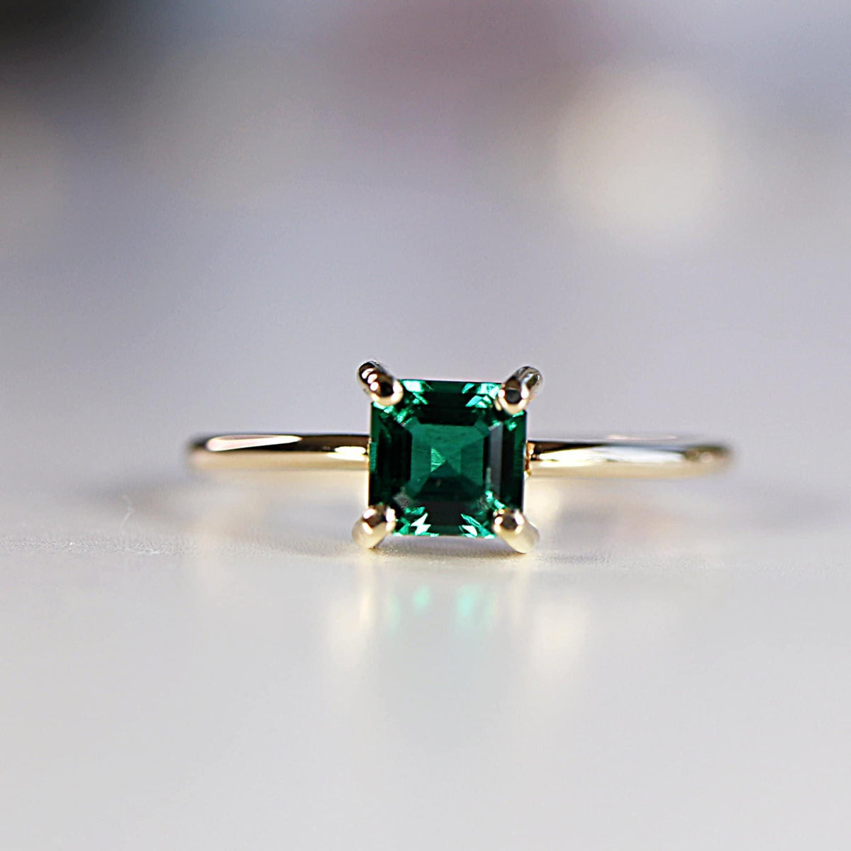 Front view of a gold ring with a square emerald-cut green gemstone set in four prongs on a plain surface.