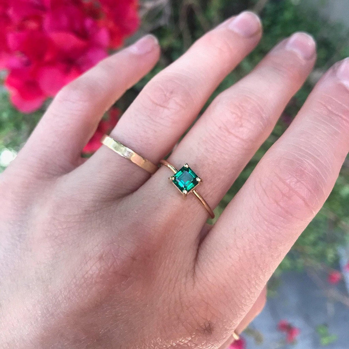 Hand wearing a thin gold ring with a small square green gemstone alongside a wider plain gold band against a background of blurred foliage.