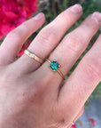 Hand wearing a thin gold ring with a small square green gemstone alongside a wider plain gold band against a background of blurred foliage.