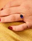 A hand with dark red nail polish wears a gold ring with an oval purple gemstone oriented horizontally, resting on a yellow textured fabric.