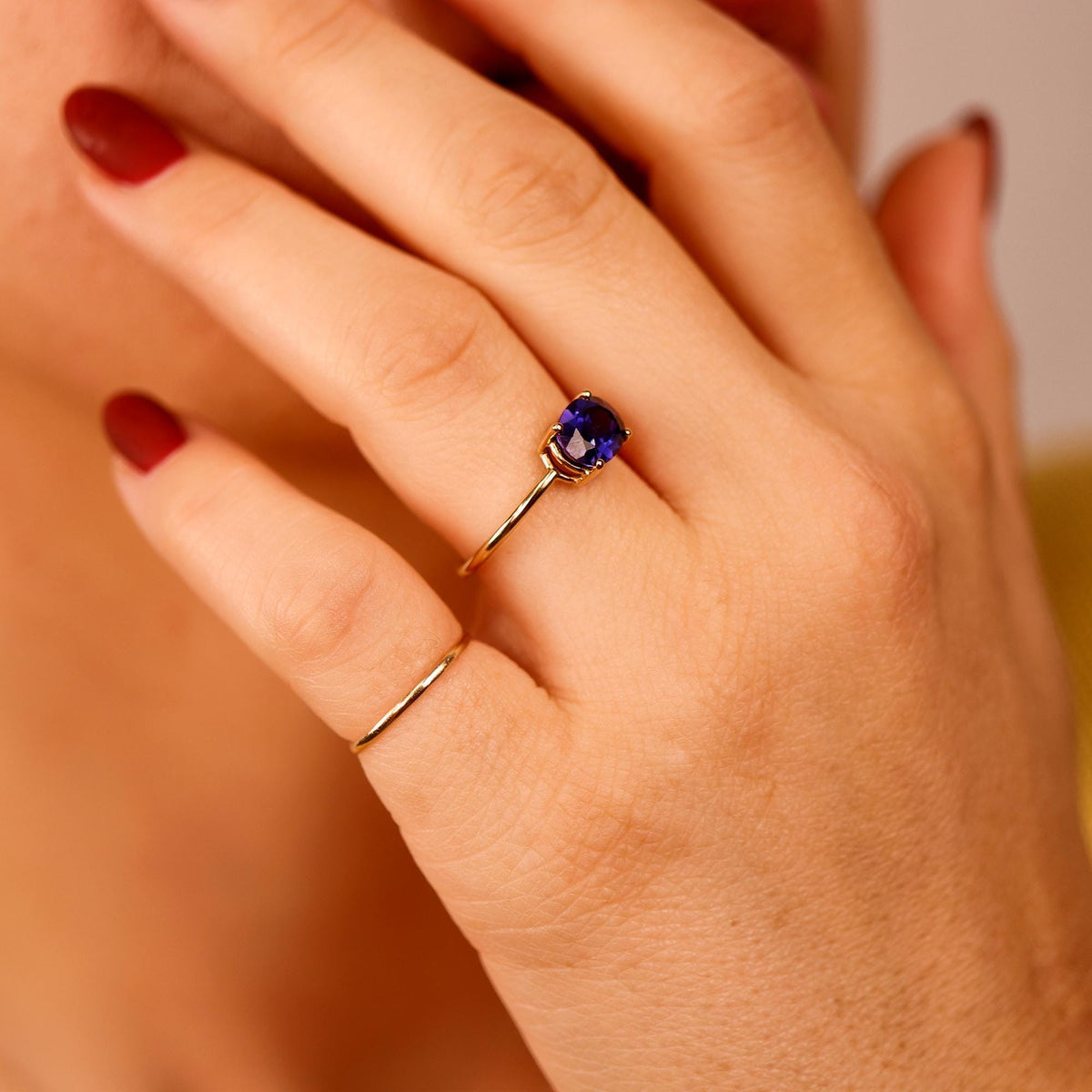 A hand wearing a thin gold band and a gold ring with a horizontal oval purple stone, positioned near the face with dark red fingernails visible.