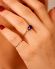 A hand wearing a thin gold band and a gold ring with a horizontal oval purple stone, positioned near the face with dark red fingernails visible.