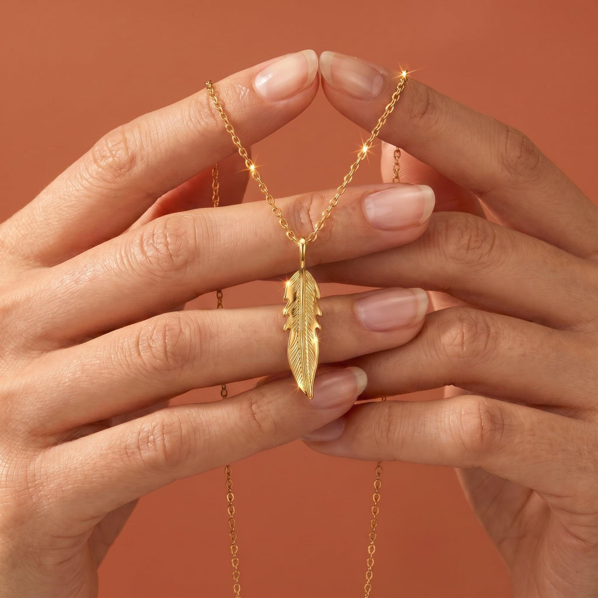 A pair of hands gently holds a gold feather pendant hanging on a thin gold chain against a solid terracotta background.