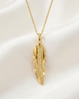 A gold feather necklace rests centrally on a soft white draped satin fabric surface.