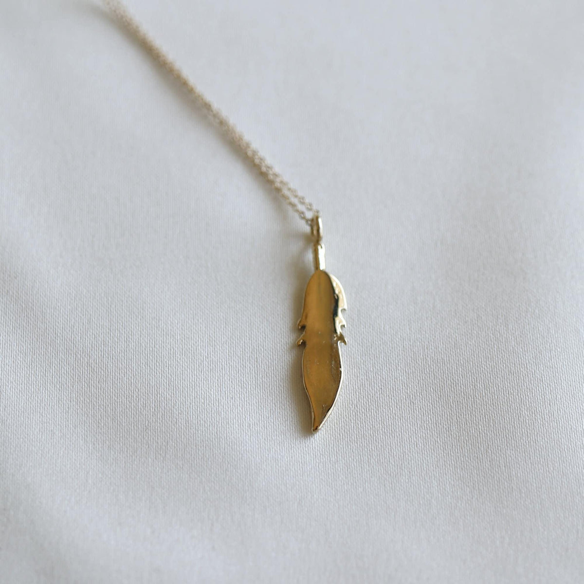 The smooth reverse side of a gold feather pendant on a fine chain lies against a white fabric background.
