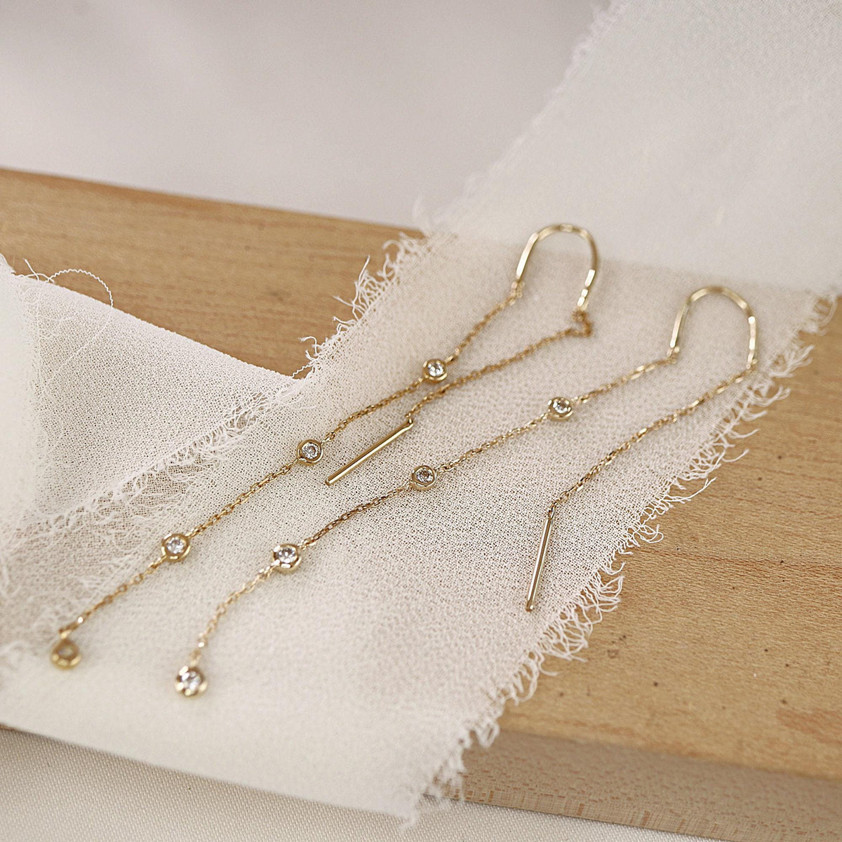 Two long gold diamond threader earrings laid across a piece of white frayed fabric and a wooden block.