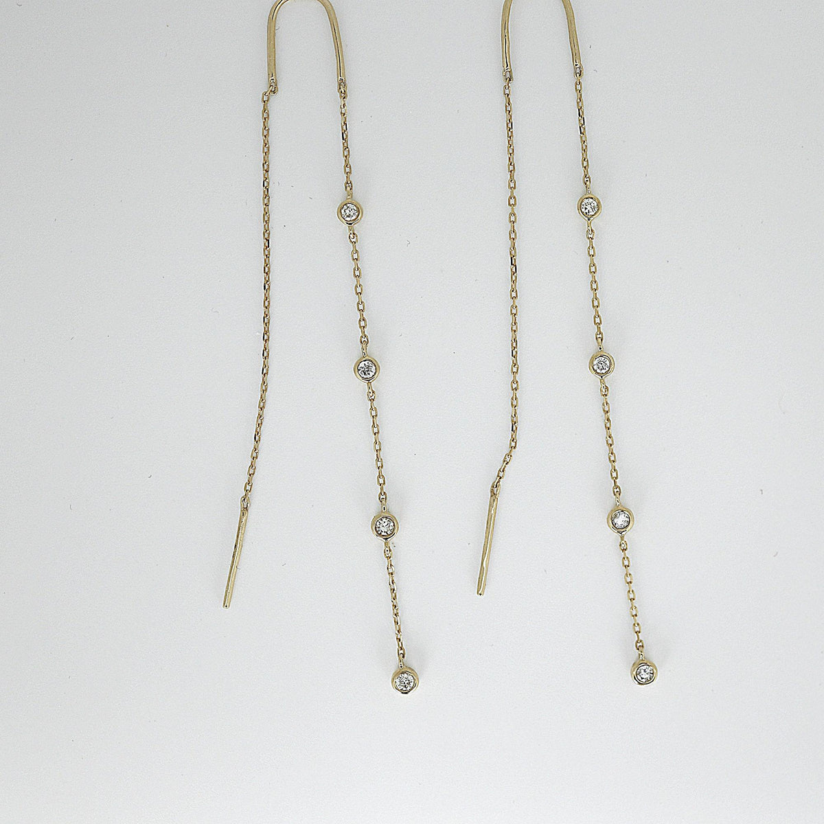Two gold threader earrings with bezel-set diamond stations laid flat on a plain white surface.
