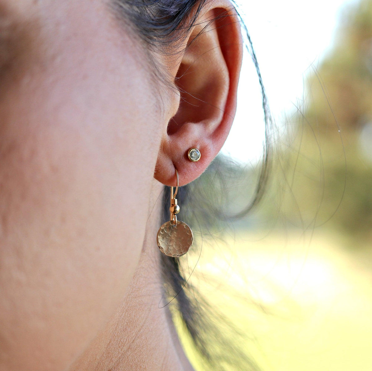 Close-up of a pierced ear wearing a gold hammered coin dangle earring and a small stud.