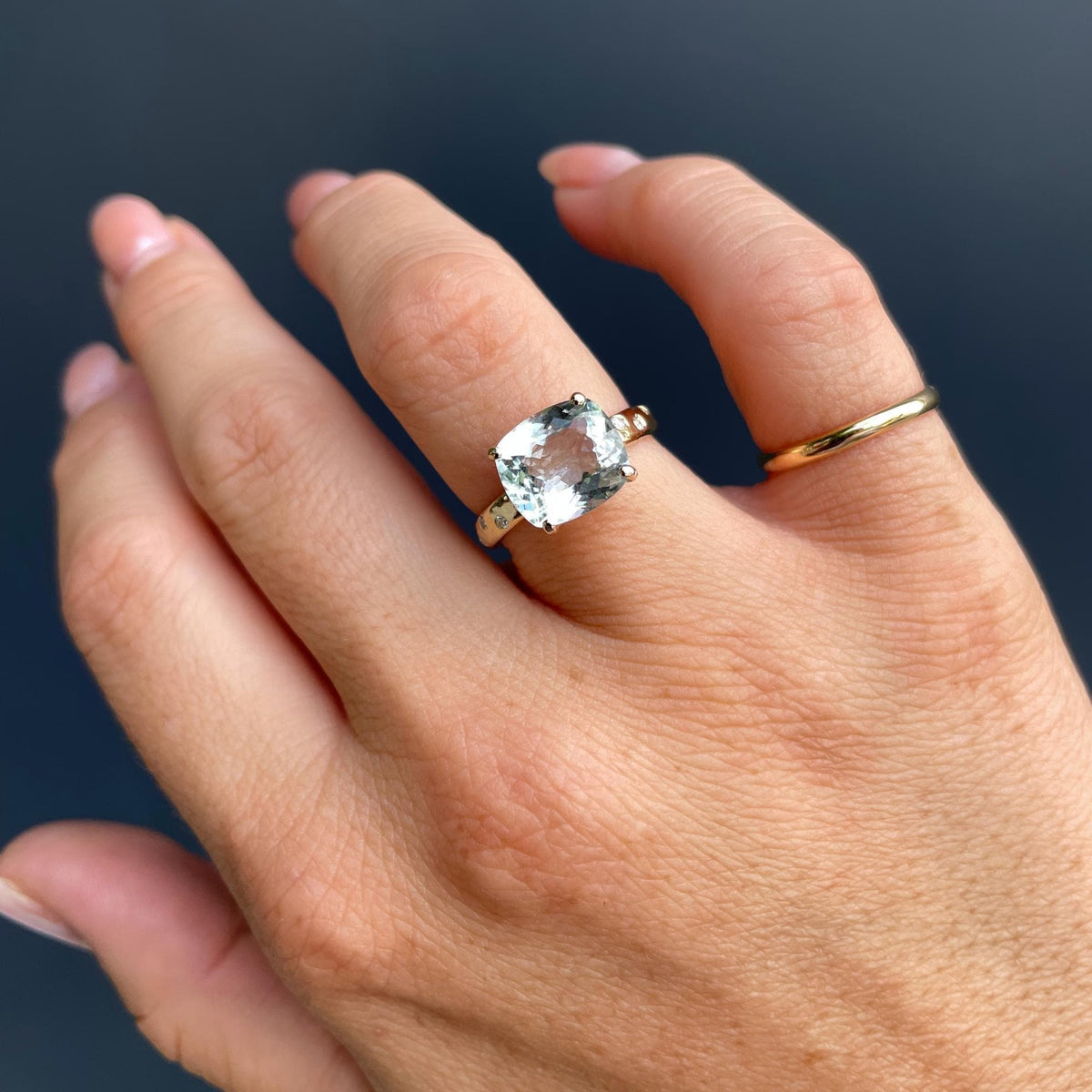 A hand wearing a gold ring with a large cushion-cut light blue gemstone and small flush-set diamonds on the band.