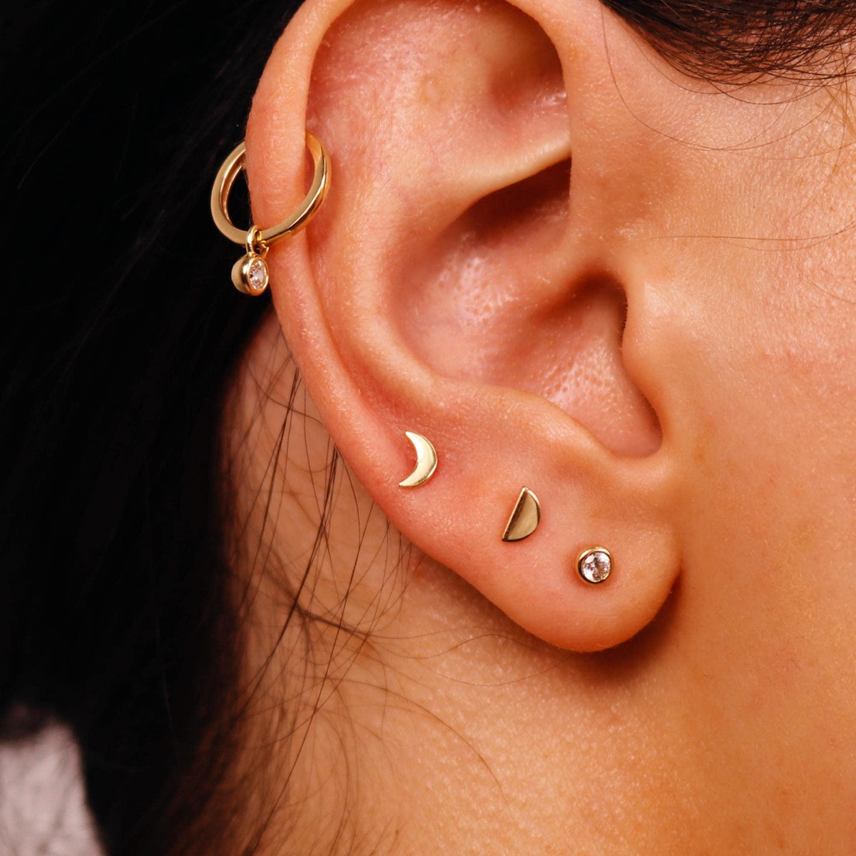 Close-up of a human ear wearing a gold hoop earring at the top, a gold crescent moon stud in the lobe, another half-moon stud, and a small round gemstone stud at the bottom.