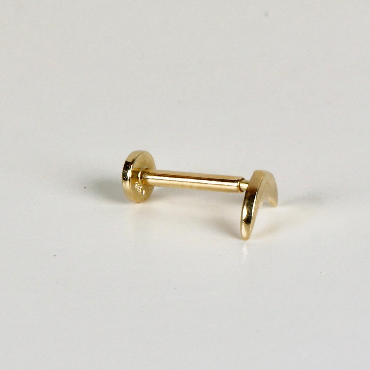 A gold flat back labret stud with a crescent moon top lying horizontally on a plain white surface.