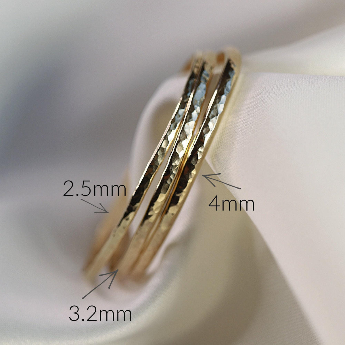 Three gold hammered cuff bracelets of varying widths resting on a white fabric surface with text labels indicating 2.5mm, 3.2mm, and 4mm.