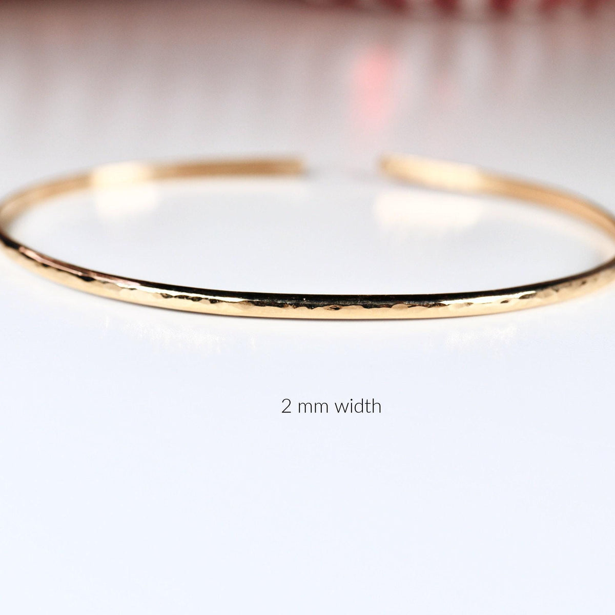 A thin gold hammered open cuff bracelet rests on a white surface with text reading 2 mm width below it.