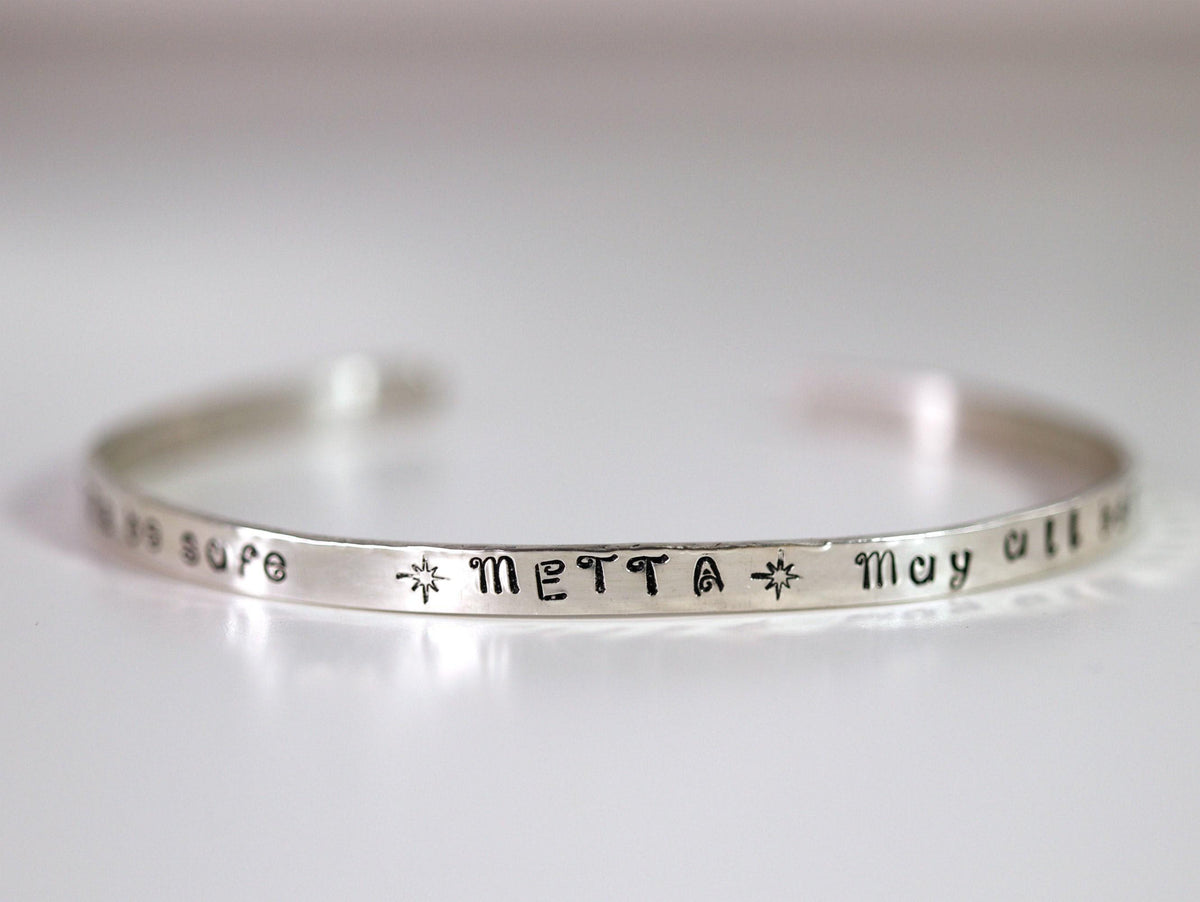 A thin silver cuff bracelet stamped with the word METTA flanked by star symbols against a plain background.