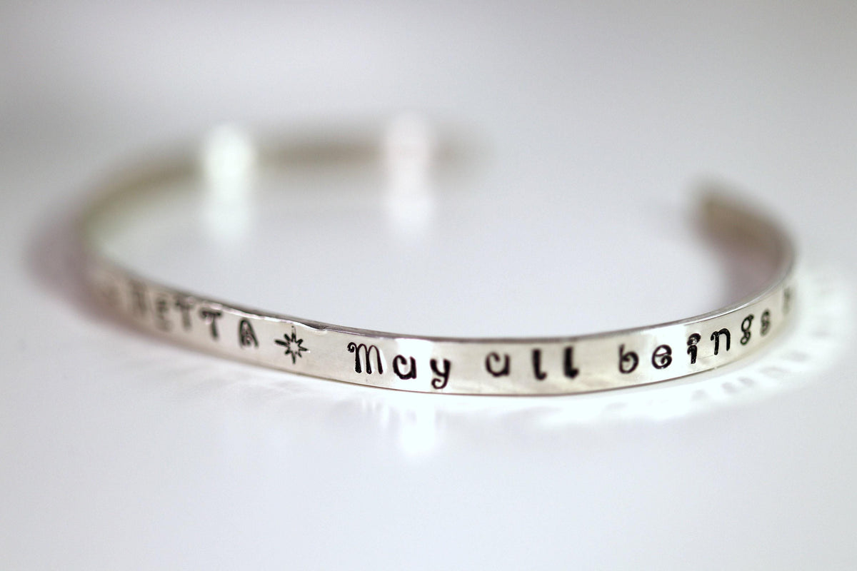 A silver cuff bracelet viewed from an angle, featuring the stamped words May all beings.