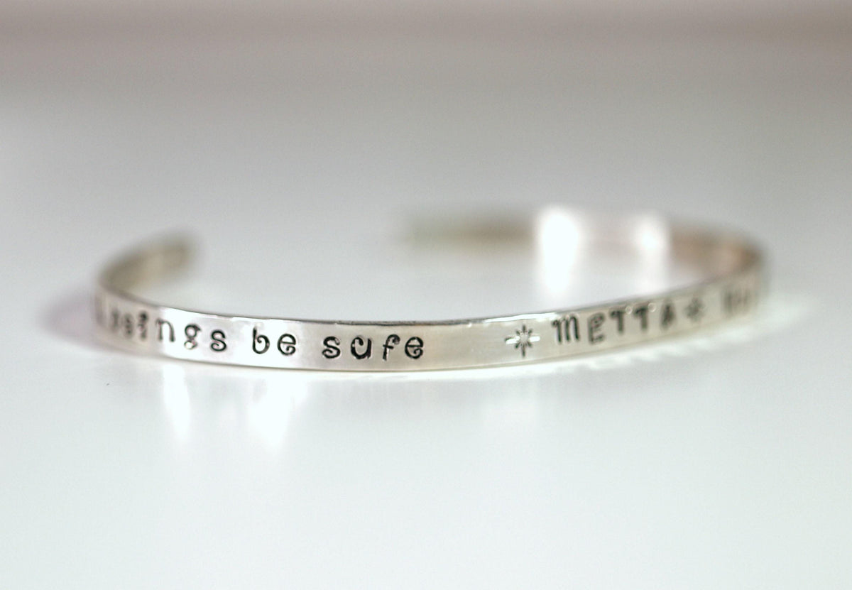 A silver cuff bracelet angled to show the stamped words be safe followed by a star and METTA.