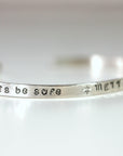 A silver cuff bracelet angled to show the stamped words be safe followed by a star and METTA.