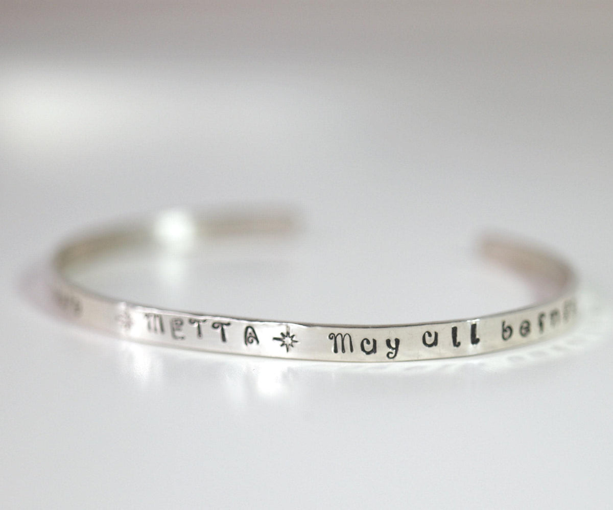A silver cuff bracelet resting on a white surface, showing the stamped text METTA and May all.