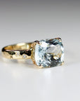 A front-facing view of the gold ring resting on a flat white surface, emphasizing the large cushion-cut light blue center stone.