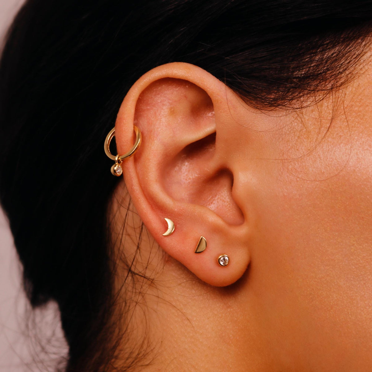Profile view of an ear adorned with a dangling charm hoop, a small crescent moon stud, a half-moon stud, and a bezel-set stone stud.
