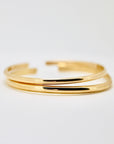 Two smooth polished gold open cuff bracelets lie stacked on a plain white background.