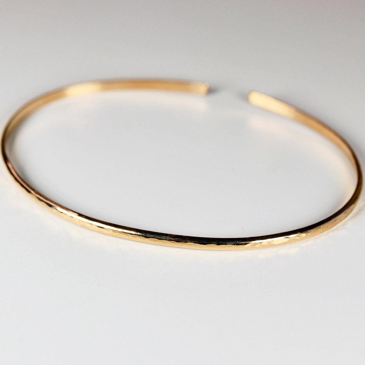 A single hammered gold open cuff bracelet rests flat on a plain white surface.