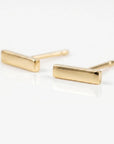 Two smooth, polished gold bar stud earrings resting on a reflective white surface.