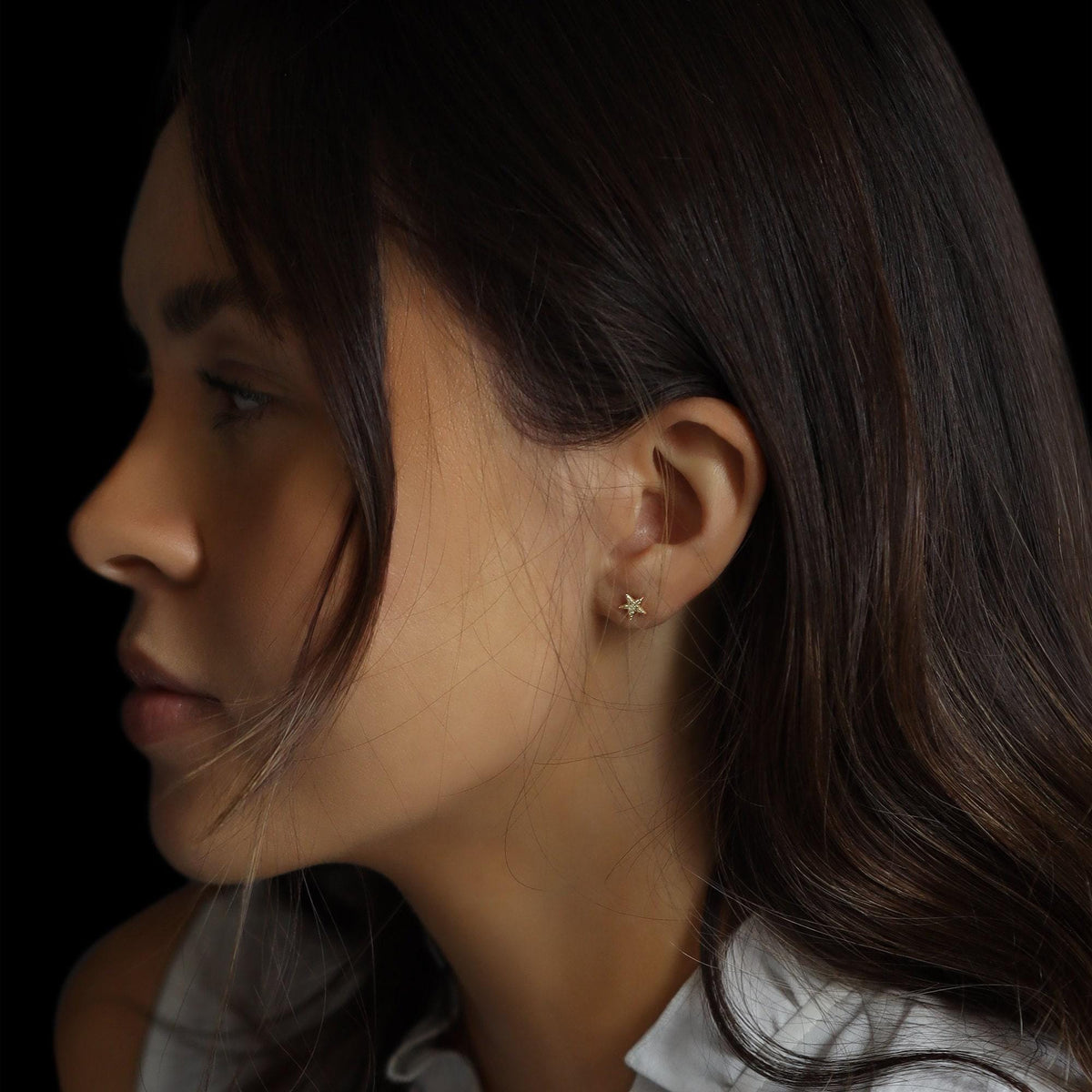 Profile view of a person with dark hair against a black background, wearing a tiny star-shaped earring.