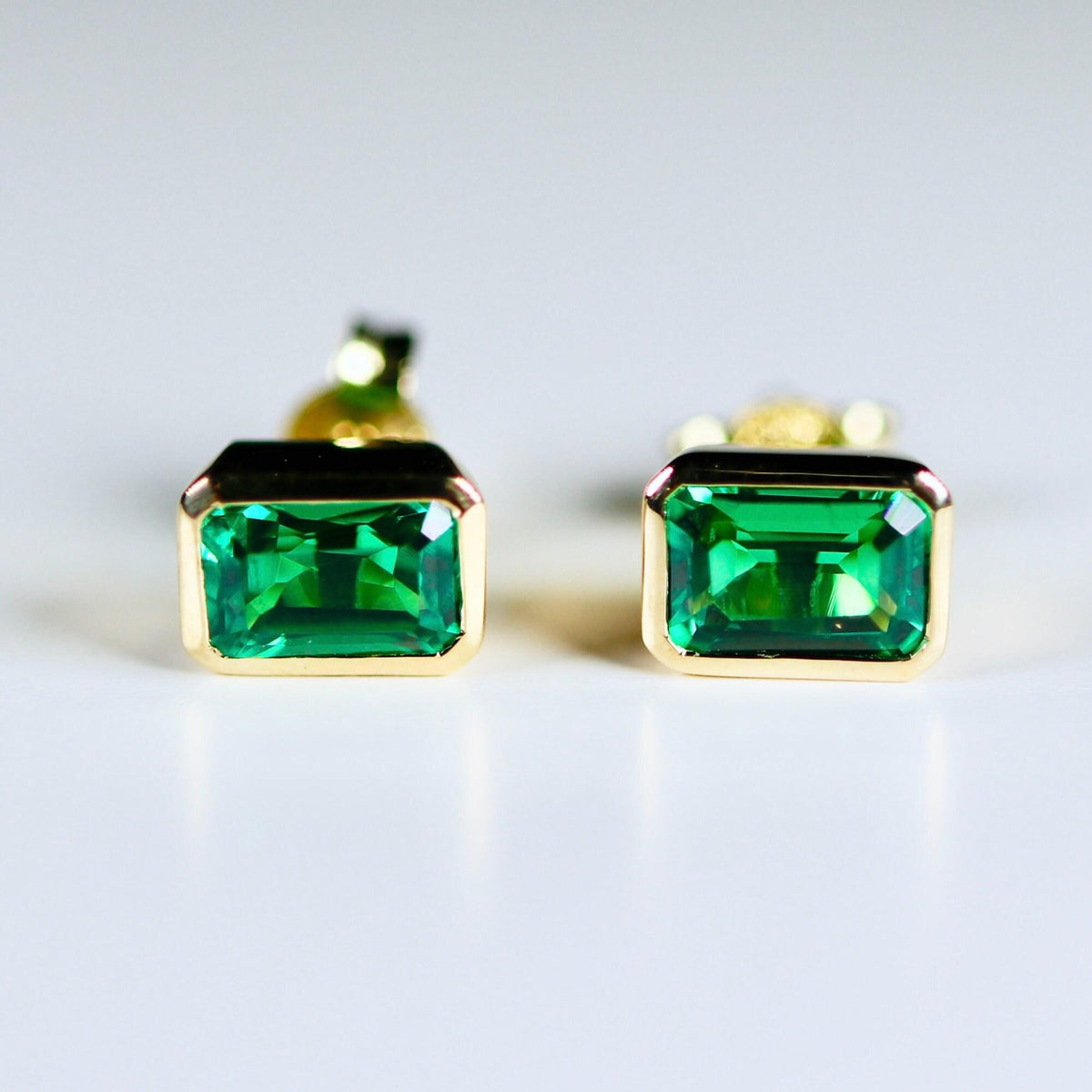 Two emerald-cut green gemstone stud earrings in yellow gold bezel settings viewed straight on against a light background.