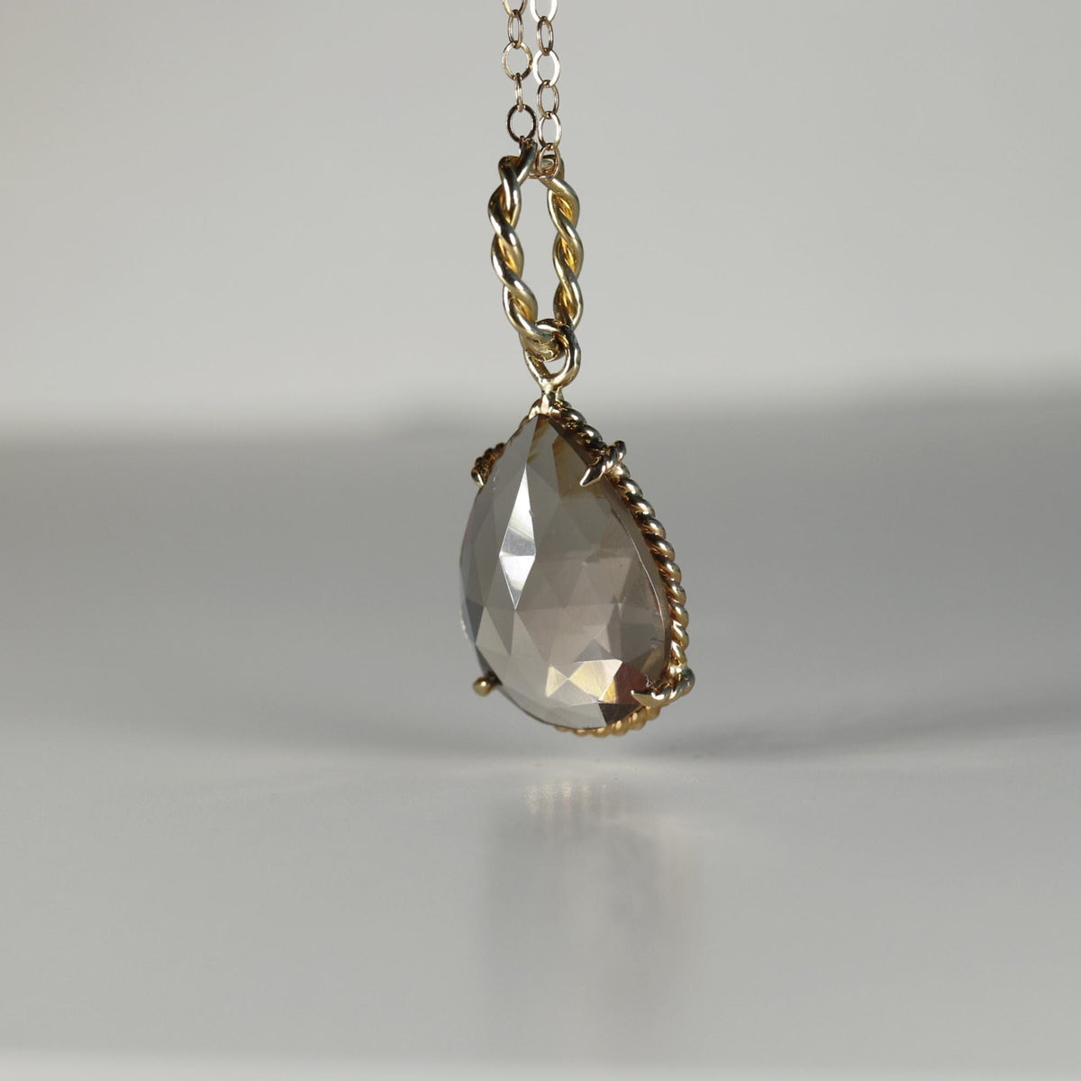 Side angle view of a pear-shaped faceted smokey quartz gemstone in a twisted gold setting suspended on a chain.