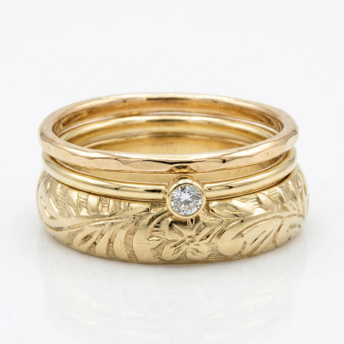 A stack of three gold rings on a white surface, featuring a wide engraved botanical band, a thin bezel-set diamond band, and a plain band.