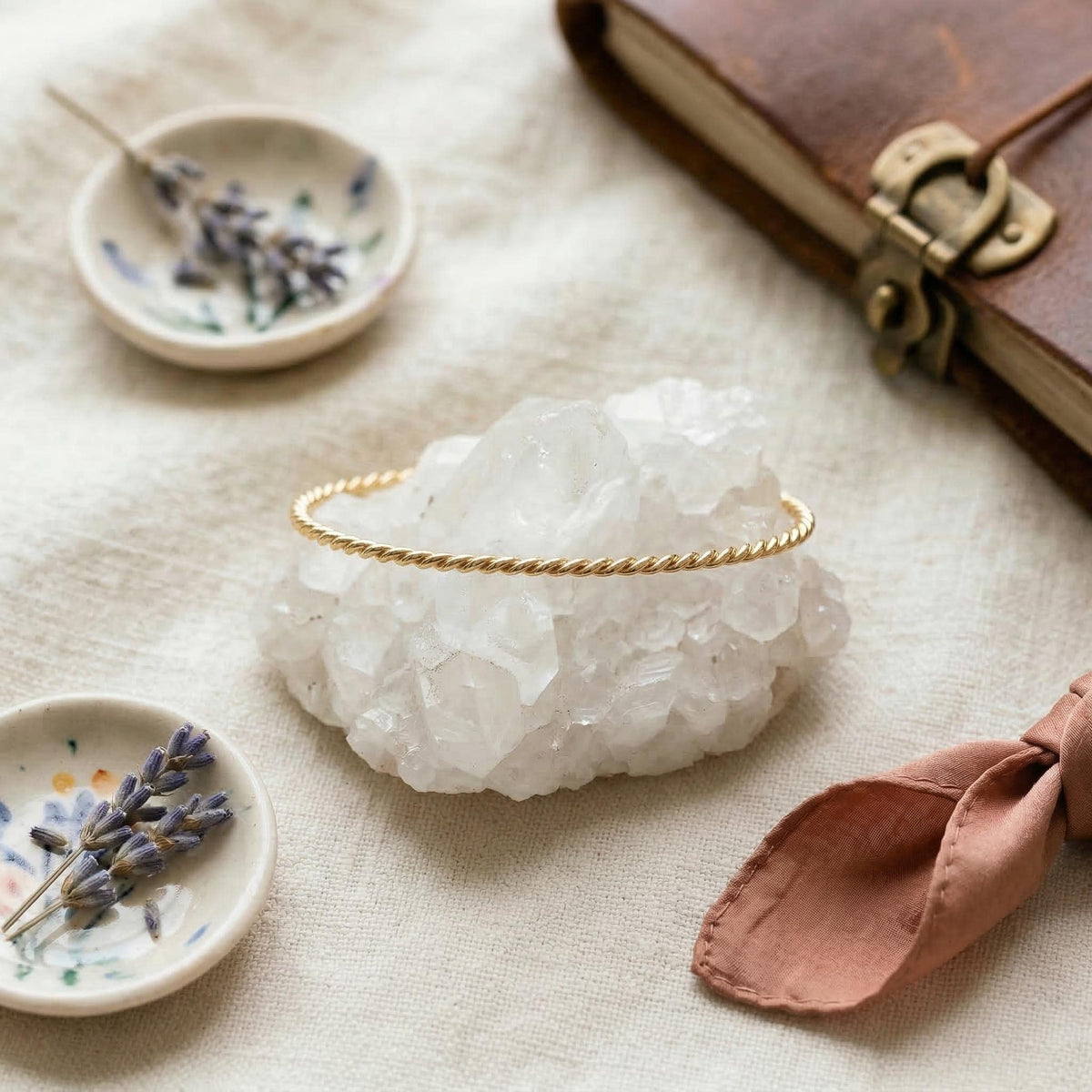 A gold twisted ring sits on a white quartz crystal next to two small bowls of dried lavender and a pink cloth.
