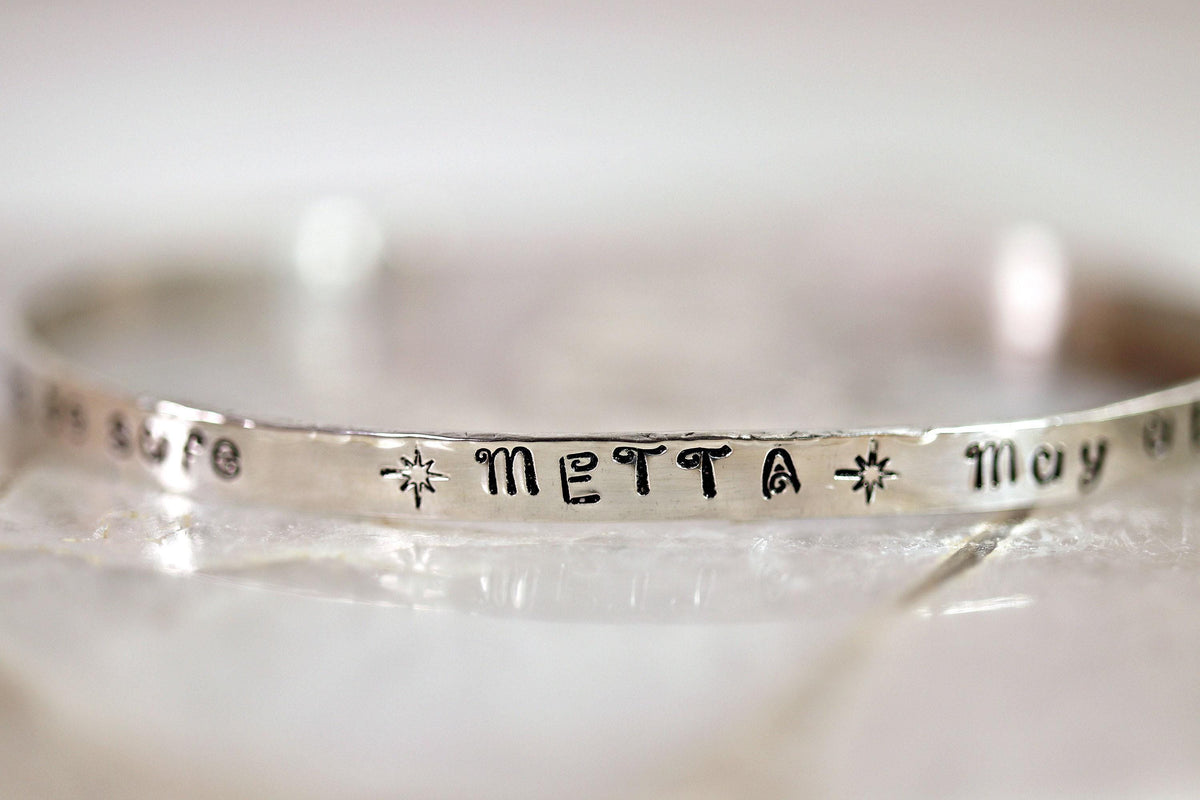 Close-up of a silver cuff bracelet resting on a textured surface, showing the stamped text METTA.
