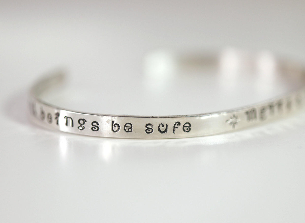 A silver metal cuff bracelet angled to display the hand-stamped phrase be safe.