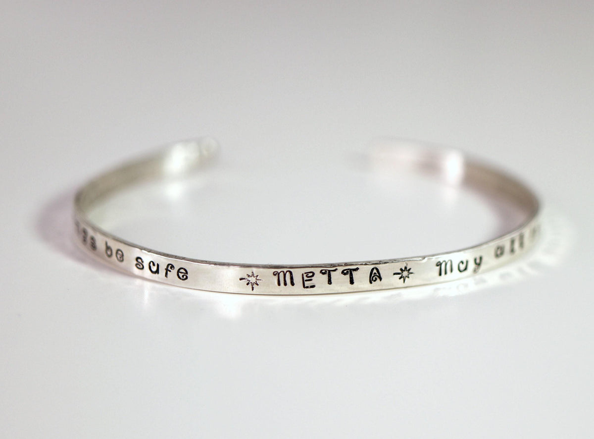 A full view of the thin silver cuff bracelet resting on a flat white surface, displaying the stamped word METTA.