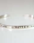 A full view of the thin silver cuff bracelet resting on a flat white surface, displaying the stamped word METTA.