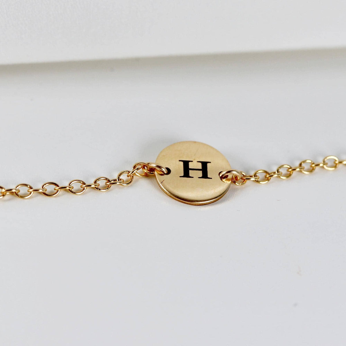 A gold chain bracelet featuring a circular disk engraved with the letter H.