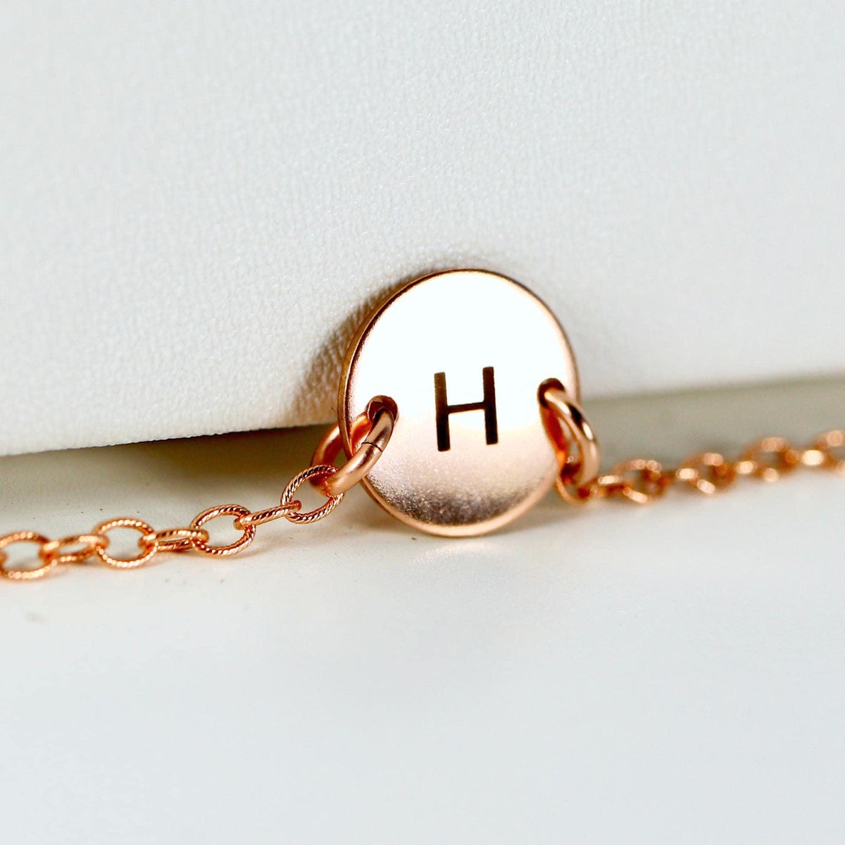 A close-up view of a rose gold circular charm engraved with the letter H on a delicate rose gold chain.