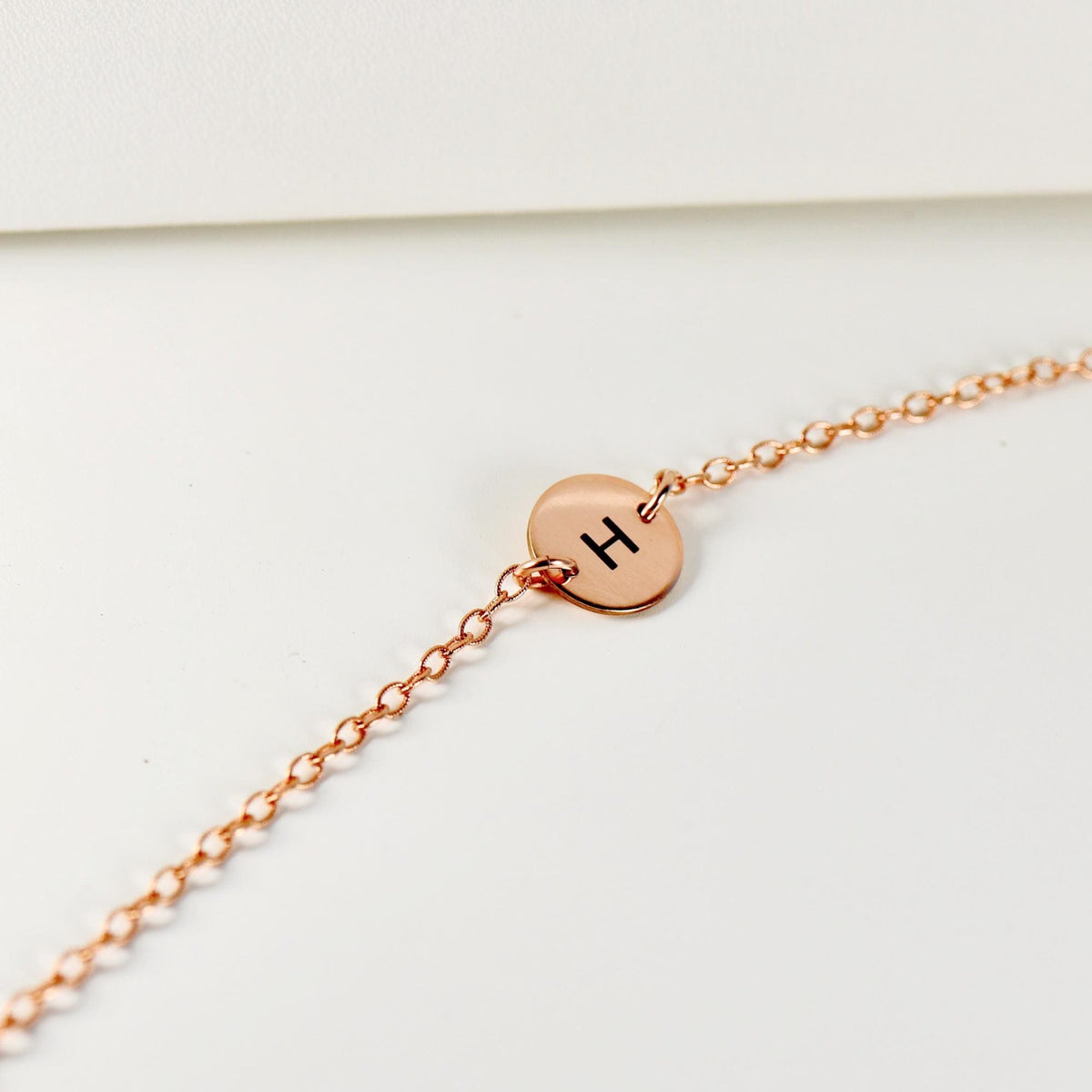 A delicate rose gold chain bracelet with a tiny round initial disk showing the letter H.