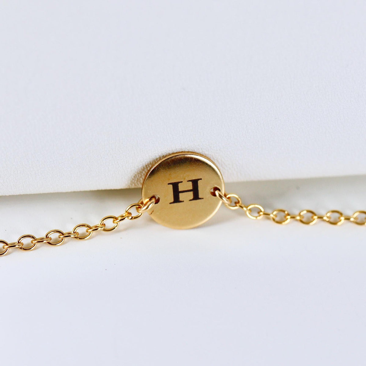 A close-up of a polished gold round disk with an engraved letter H connected to a link chain.