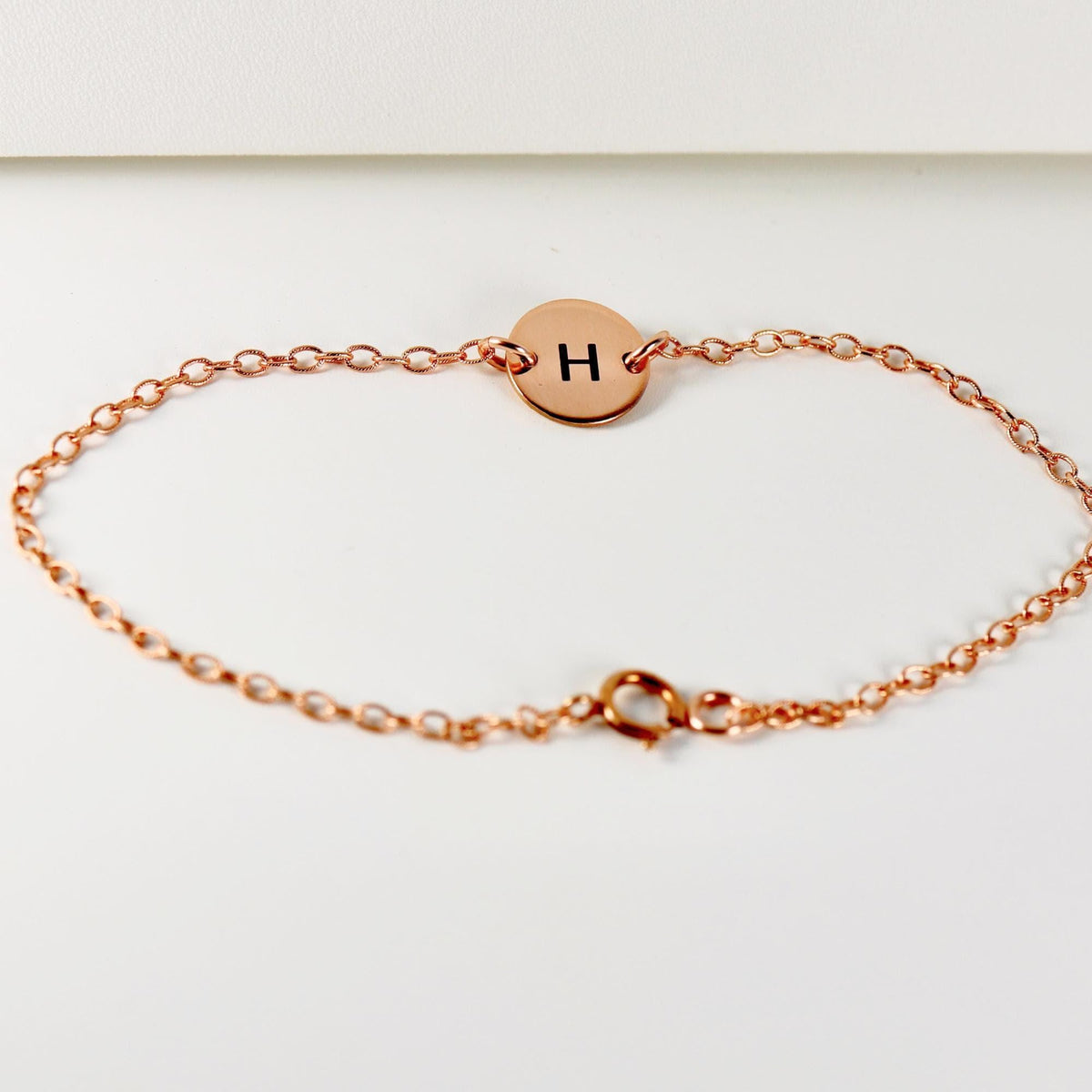 A full view of a rose gold link bracelet with a centered circular disk charm engraved with an H and a spring-ring clasp.