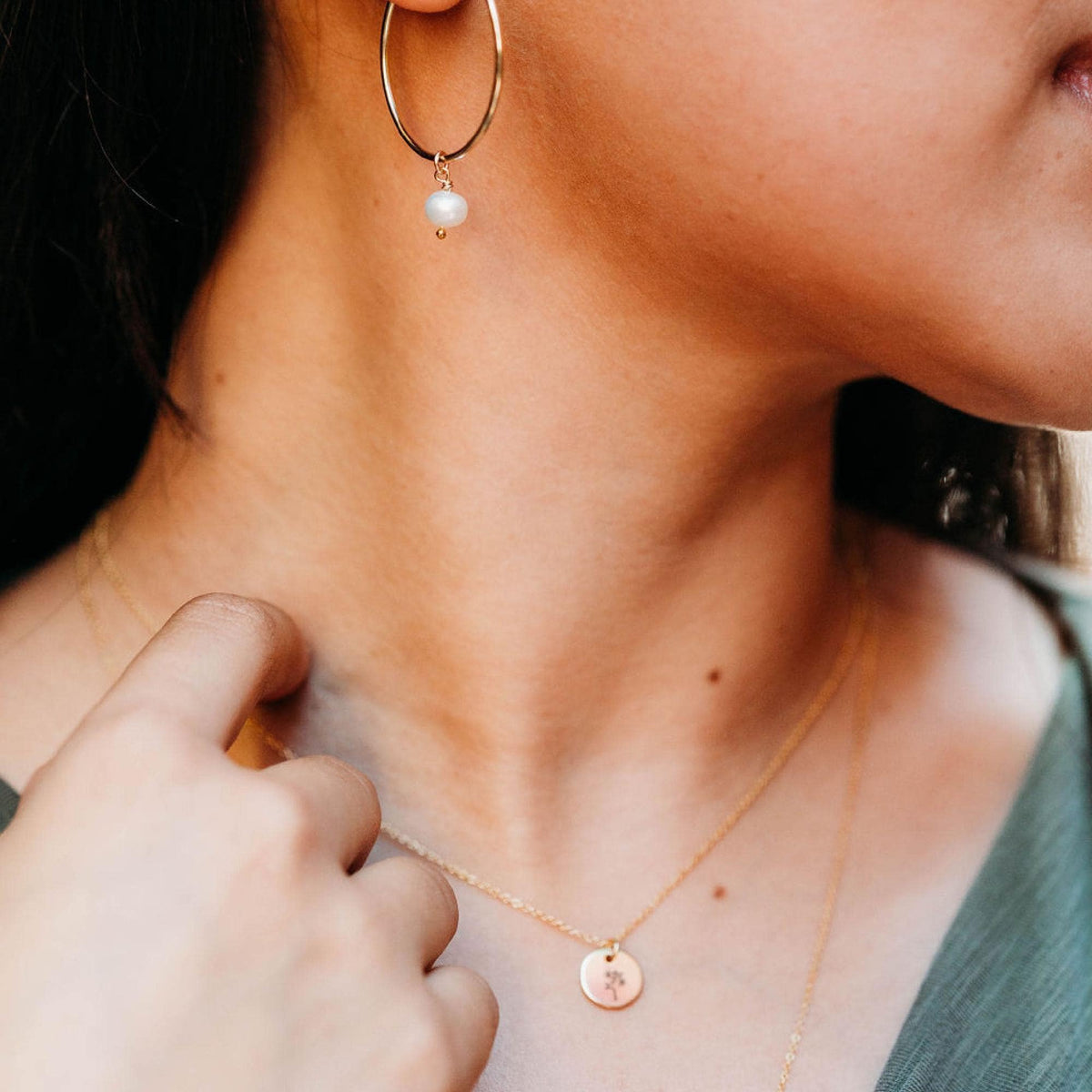 A close-up of a person touching one of two gold layered necklaces featuring engraved disc pendants.
