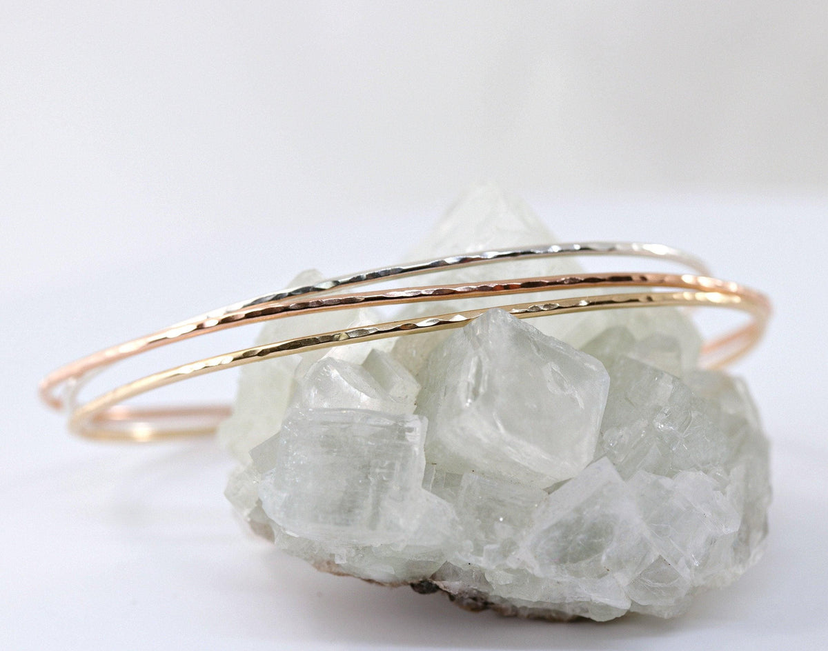Three thin hammered metal cuff bracelets in silver, rose gold, and yellow gold rest on a rough crystal cluster.
