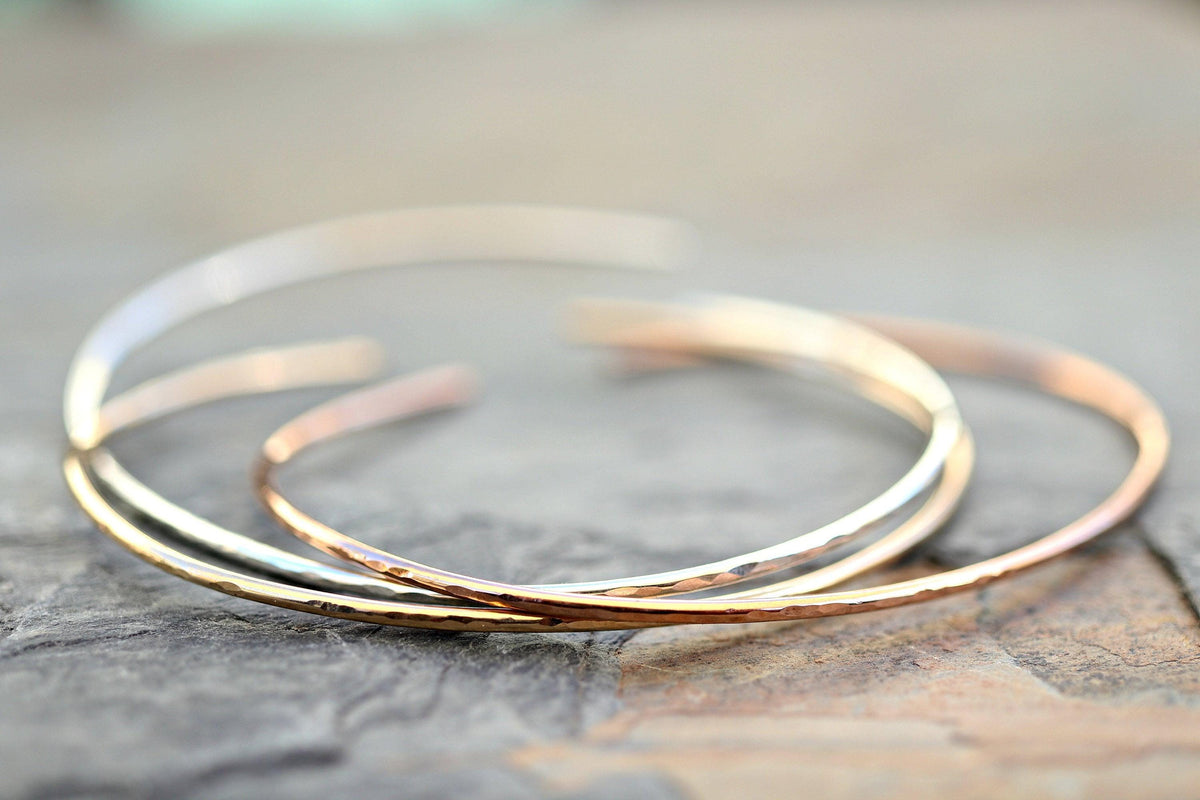 Three thin metal open cuff bracelets in silver, rose gold, and yellow gold are stacked on a rough gray surface.