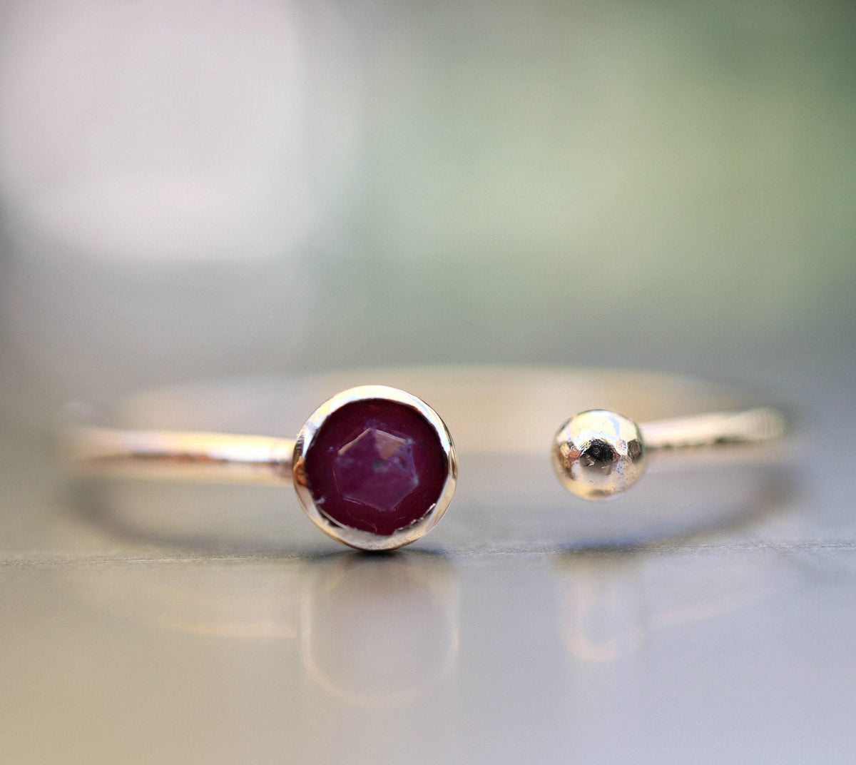 Low-angle shot of the gold ring on a reflective surface, highlighting the hexagonal rose-cut ruby and spherical ball terminal.