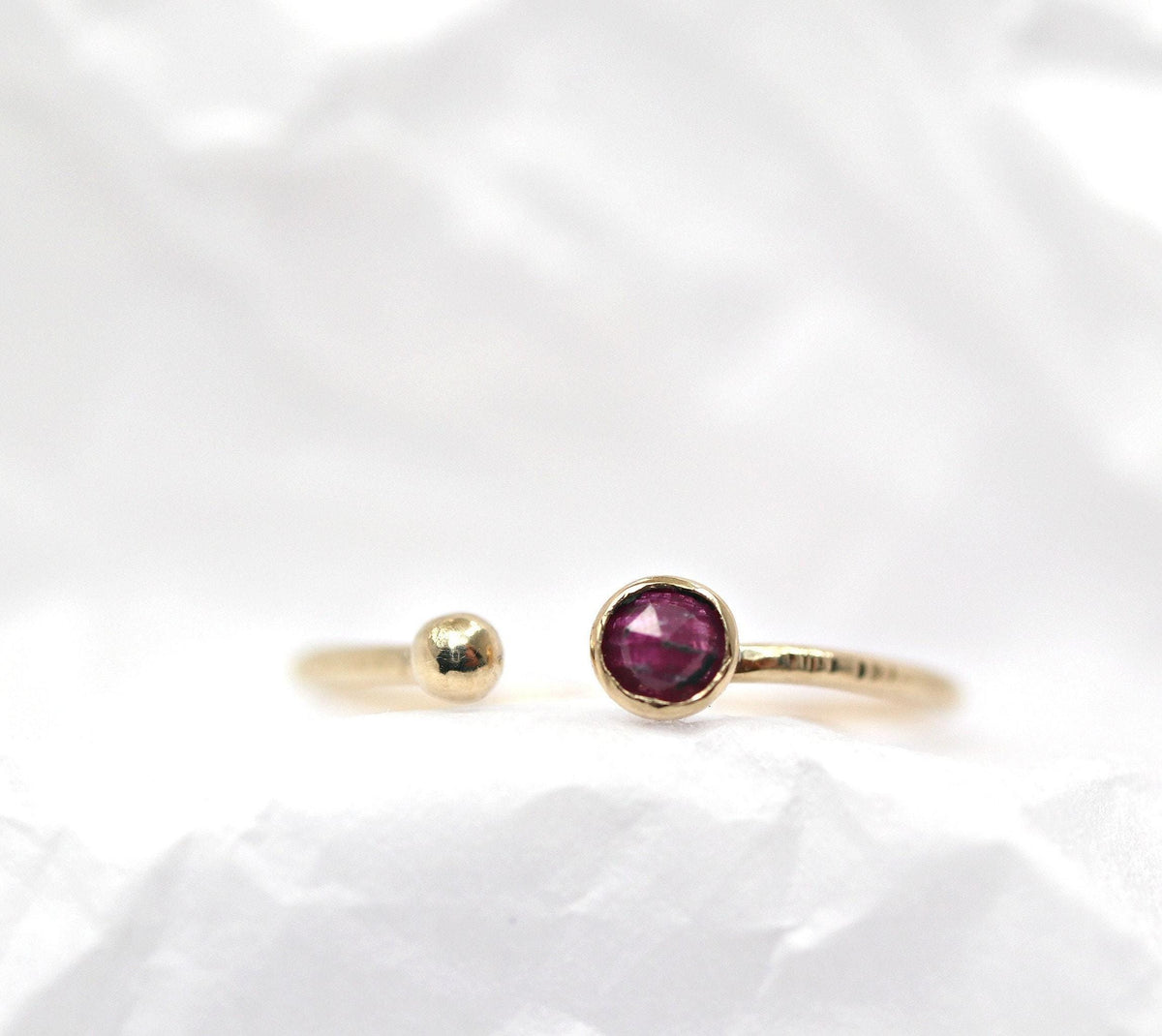 Frontal view of the gold open ring with a textured band, featuring a round ruby and a ball terminal against a white crinkled background.